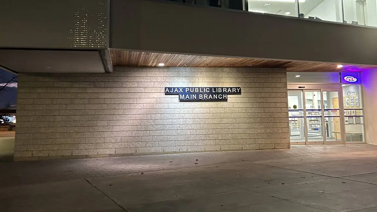 Night exterior of the Ajax Public Library main branch entrance and empty paved plaza; no people visible.