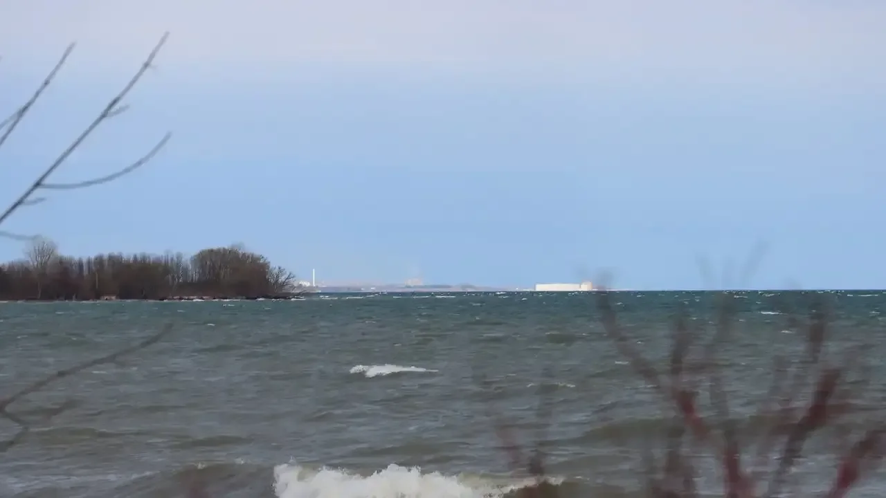Lake Ontario shoreline in Ajax looking east, with waves, bare trees and the Darlington Nuclear Plant on the horizon.