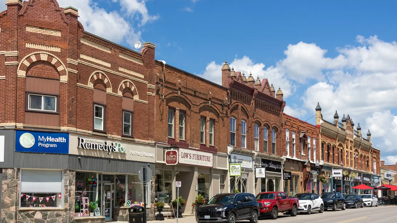 Historic brick storefronts on Queen Street in downtown Port Perry, Ontario, with parked cars under a blue sky; no people visible.