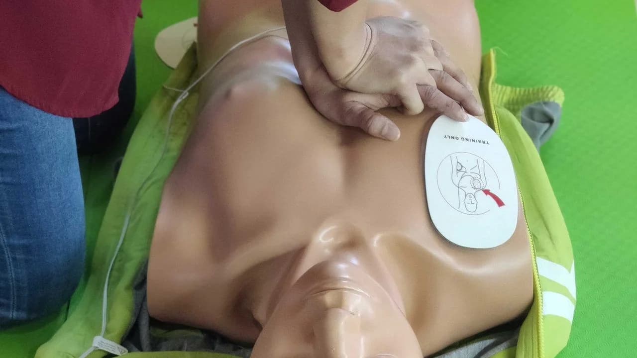 Trainer's hands practicing chest compressions on a CPR training mannequin lying on a green exercise mat; no real people visible