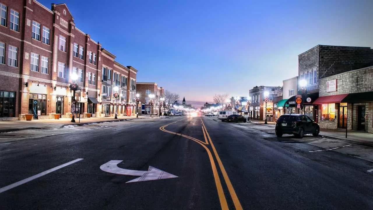 Downtown main street at dusk with brick storefronts, glowing street lamps and parked cars; no pedestrians.