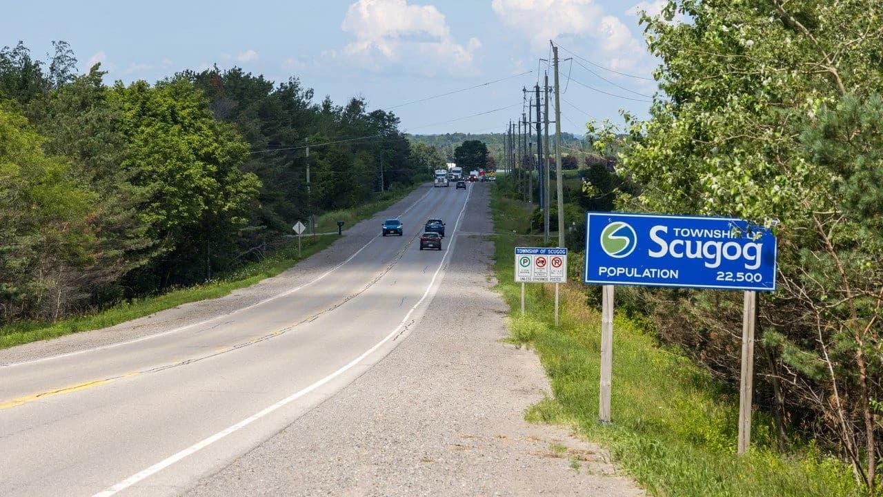 Daytime view of a rural two-lane road entering the Township of Scugog, Ontario, with a Township of Scugog sign; no people visible.