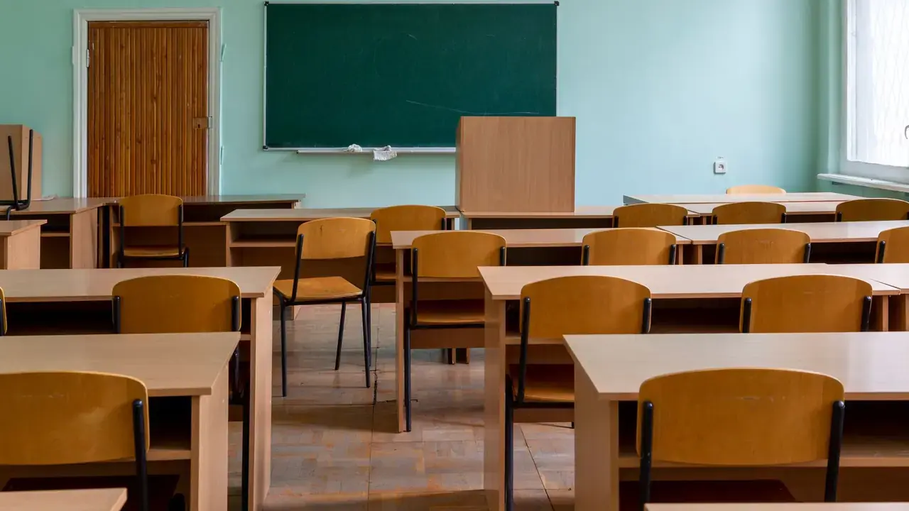 An empty classroom with rows of wooden desks and chairs facing a blank green chalkboard; no people visible.