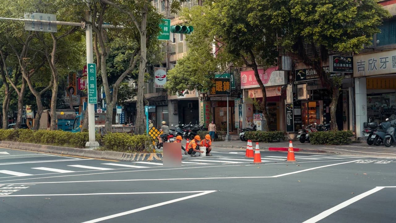 Road workers in orange vests painting lane markings at a city intersection with traffic cones.