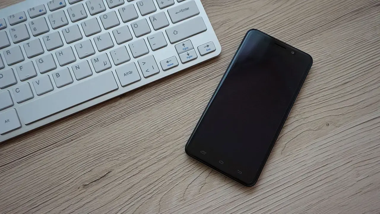 A black smartphone with a blank dark screen beside a keyboard on a wooden desk; no people visible.