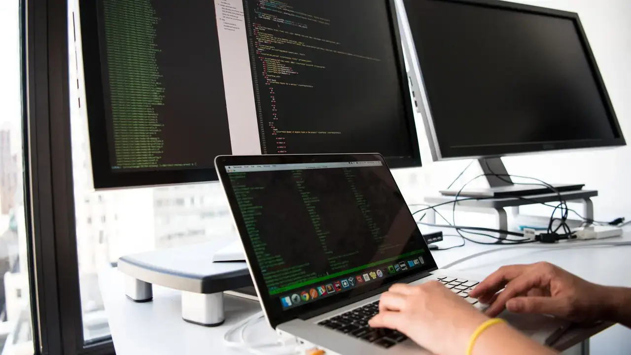 Hands typing on a laptop in front of monitors showing code; no face visible.