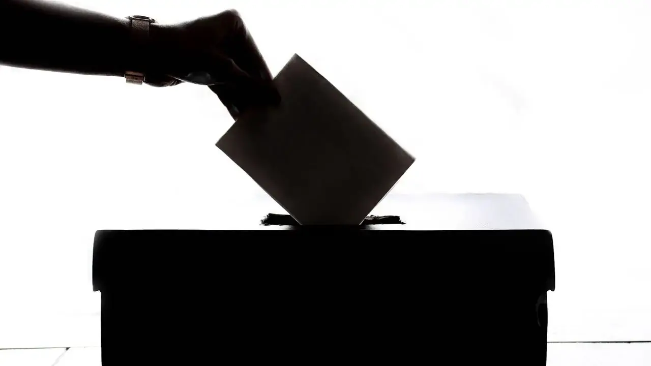 Silhouette of a hand placing a ballot into a ballot box; no faces visible.