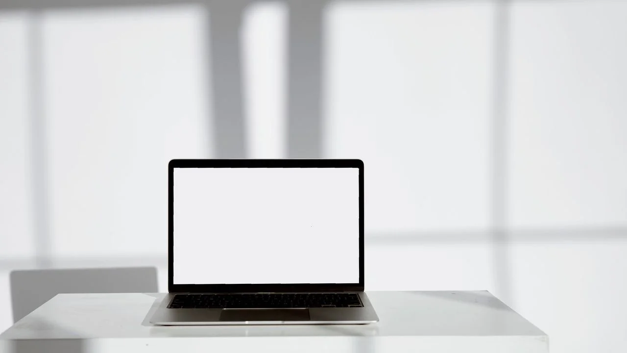 Grayscale photo of a laptop with a blank white screen on a white desk in a sunlit room; no people.